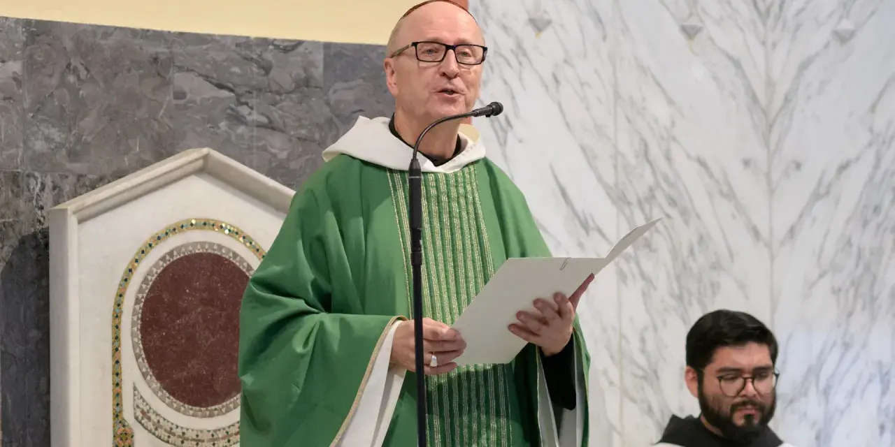 A man with a Benedictine title, wearing a green chasuble, speaks into a microphone while holding a paper.