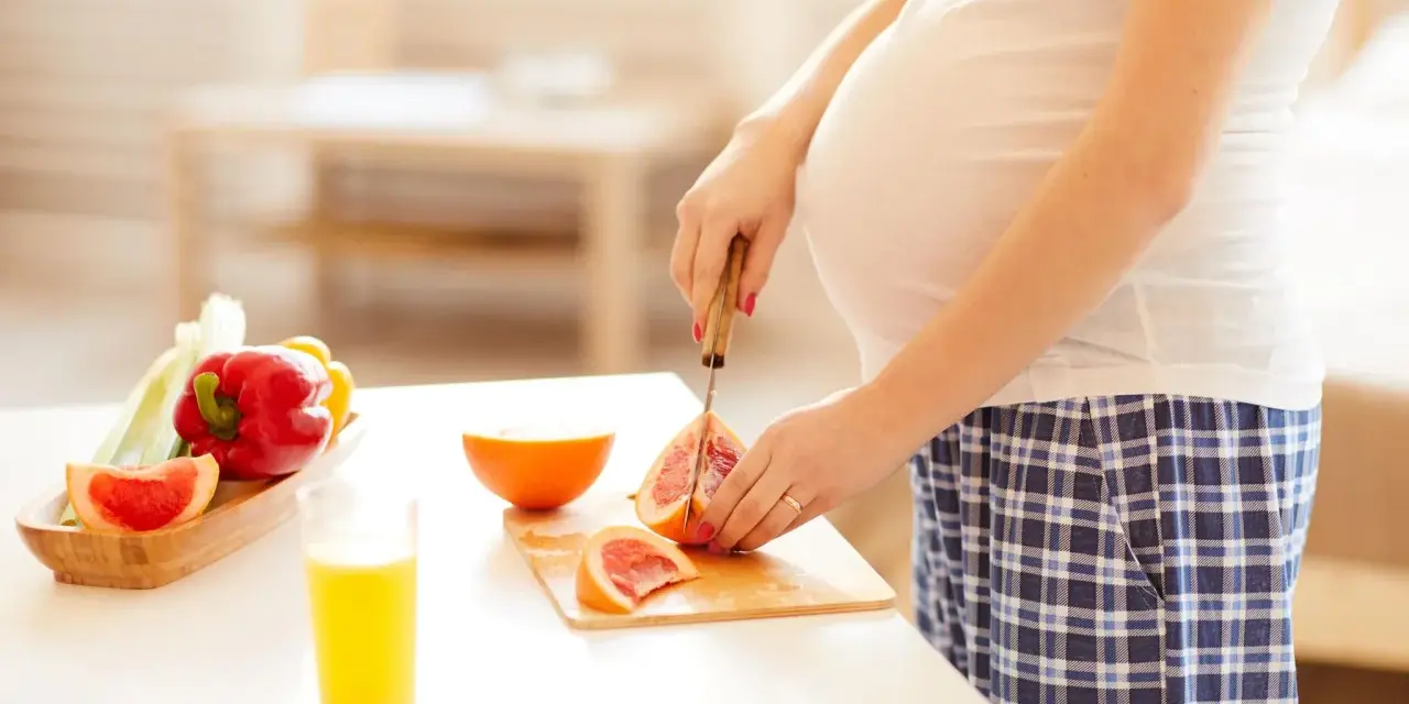 Schwanger, die Grapefruit für einen gesunden Snack schneidet. Nebenan liegen Paprika und ein Glas Saft.