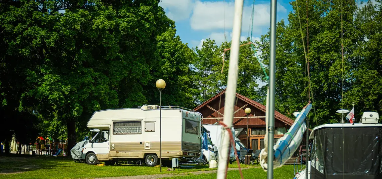 Słoneczny dzień na kempingu na Mazurach. Kamper stoi na trawie obok drewnianego budynku, w tle zielone drzewa i plac zabaw.