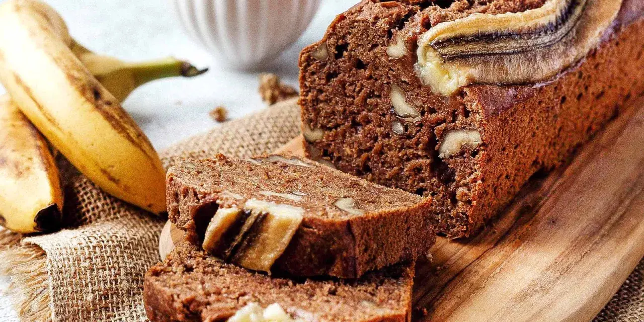 Saftiges, veganes Bananenbrot mit Walnüssen und Bananenscheiben, daneben frische Bananen und eine Schüssel Nüsse.