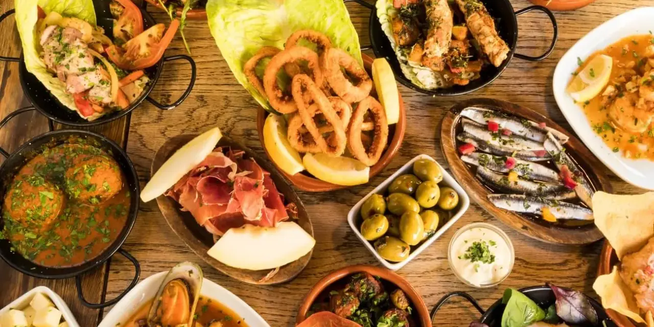 Tapas selber machen: Eine Auswahl an köstlichen spanischen Kleinigkeiten wie frittierte Tintenfischringe, Sardinen, Oliven und Schinken auf einem Holztisch.