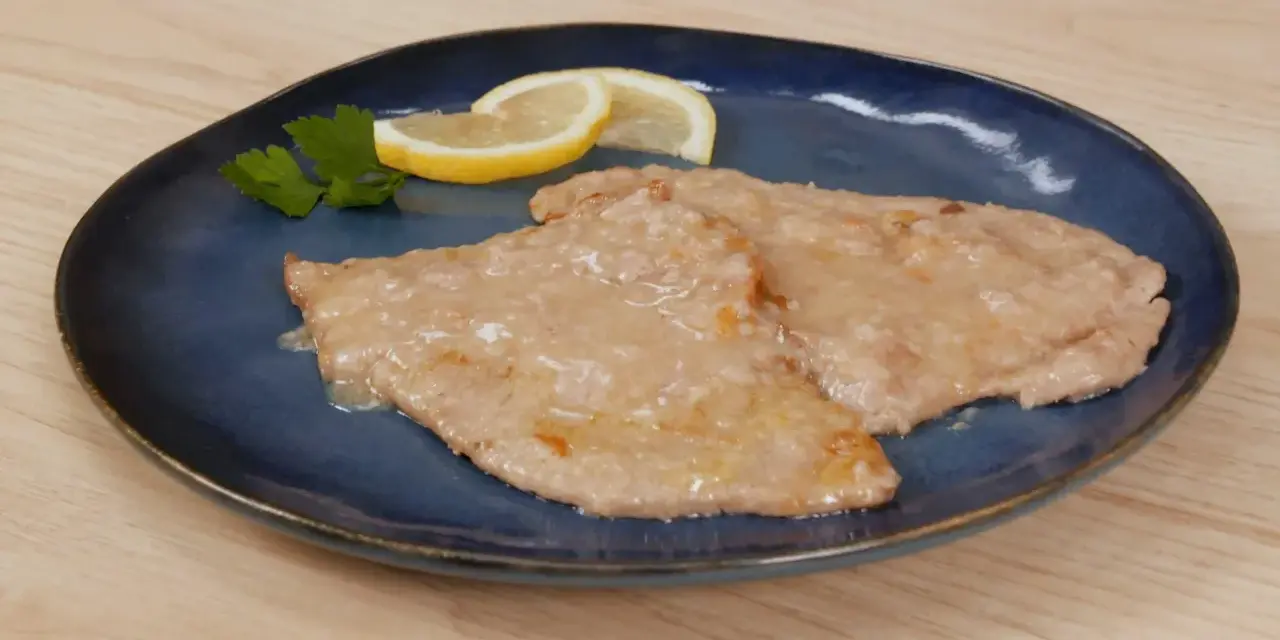 Due scaloppine al limone cremose, guarnite con prezzemolo e spicchi di limone, su un piatto blu.