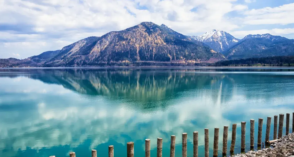 Spiegelglatter See mit Bergen im Hintergrund, wie man ihn in Bayern sehen kann. Holzpfähle ragen aus dem türkisfarbenen Wasser.