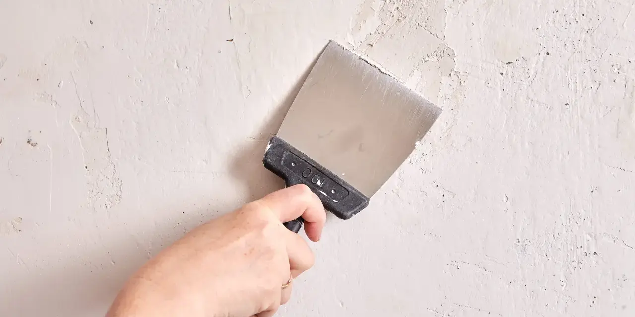 Mano con espátula metálica alisando una pared de yeso con textura irregular.