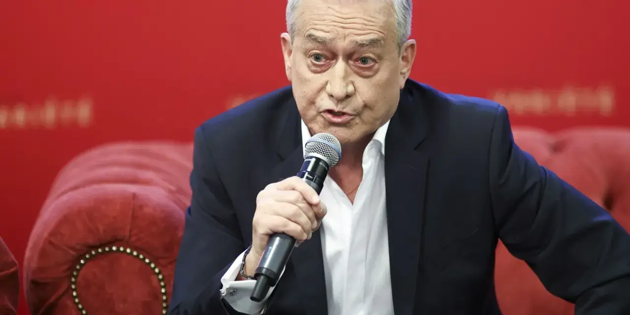 Janusz Szydłowski przemawia do mikrofonu, siedząc na czerwonej, pikowanej kanapie.