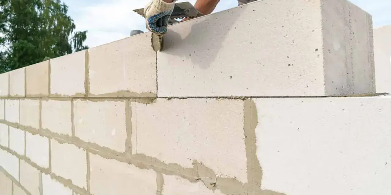 Budowanie ściany z bloczków betonowych. Ręka w rękawicy nakłada zaprawę na kolejny element.