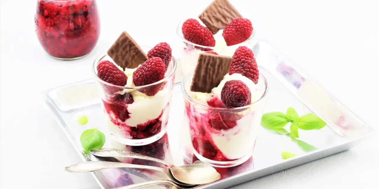 Drei Gläser mit cremigem Dessert, Himbeeren und Schokolade. Ein Glas mit Himbeermarmelade daneben. Schnelles Eis für den Sommer.