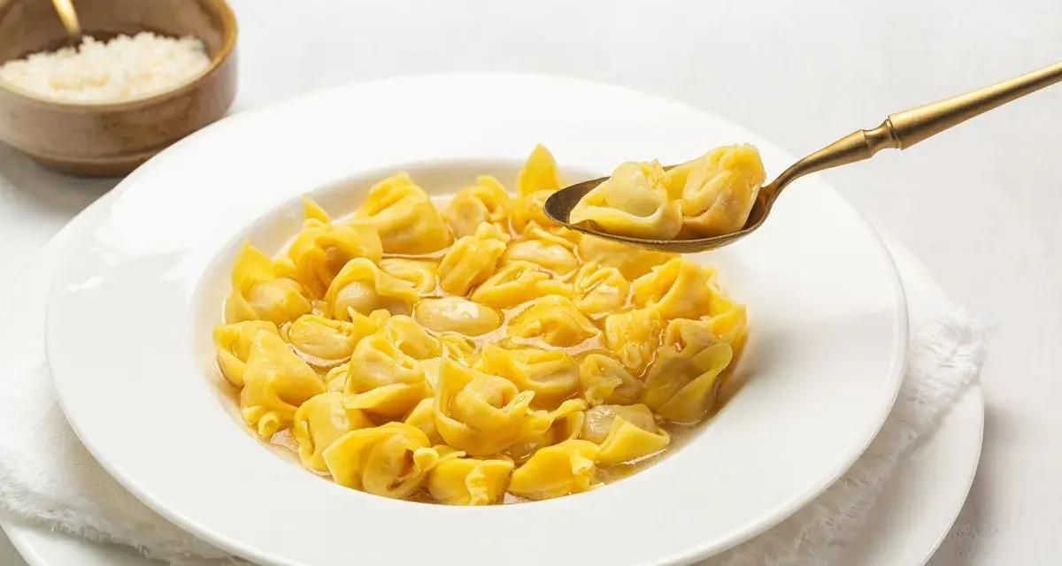 Un cucchiaio d'oro solleva un boccone di cappelletti in brodo fumante da un piatto bianco.