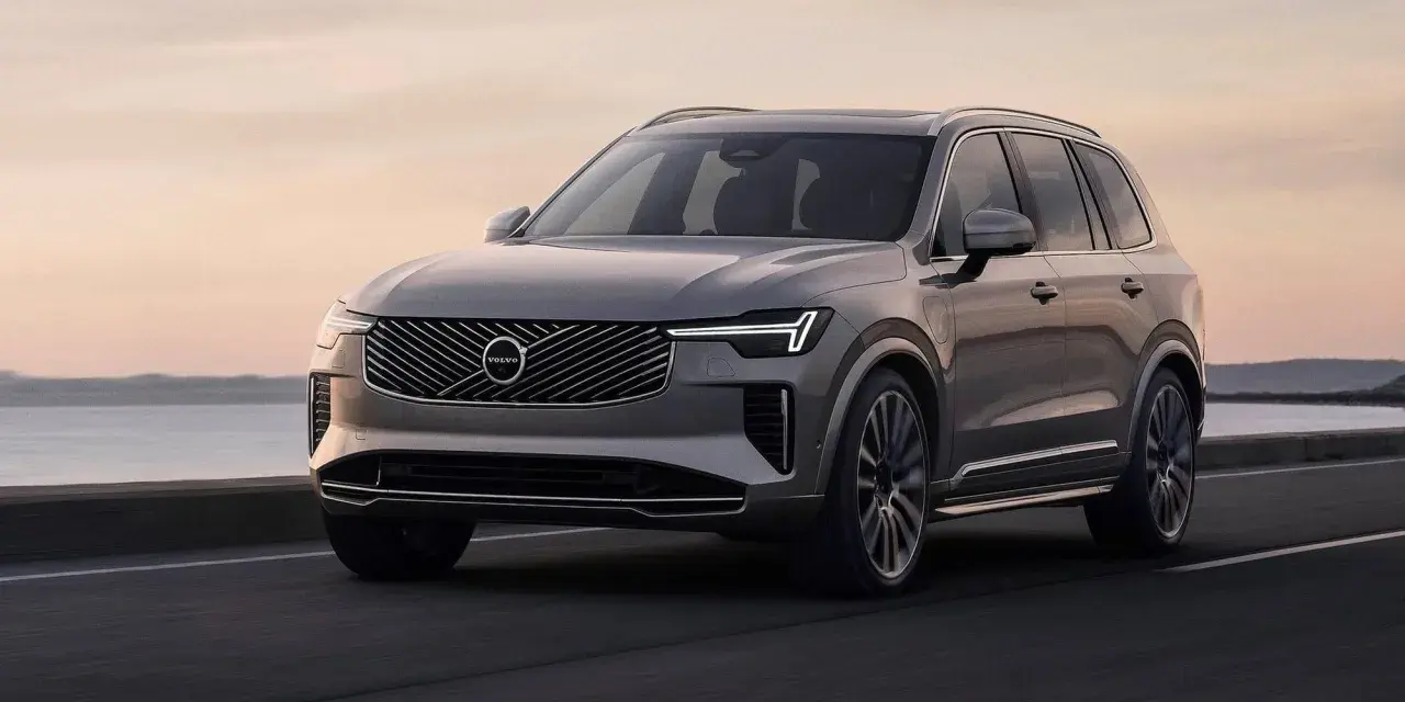Elegancki, szary Volvo CX90 jedzie po drodze nad jeziorem o zachodzie słońca.