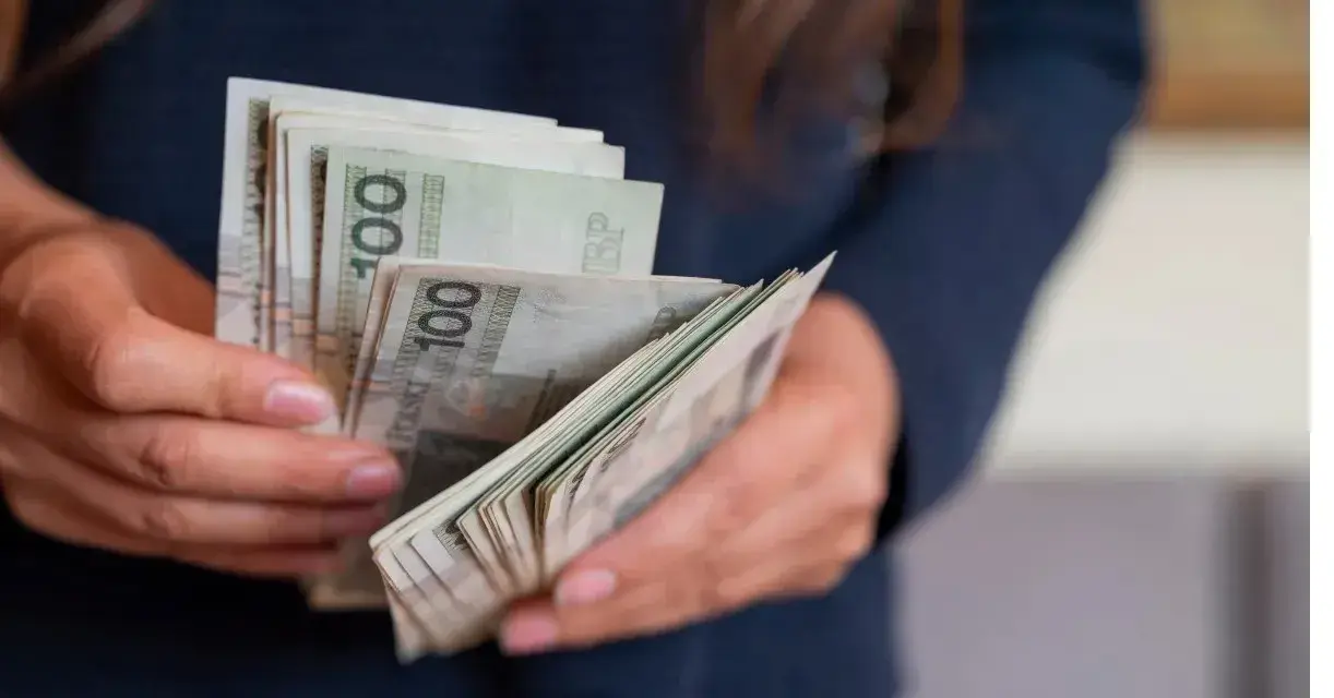 Dłonie trzymające plik polskich banknotów 100 zł. Zastanawiasz się, urlop wychowawczy ile płatny?