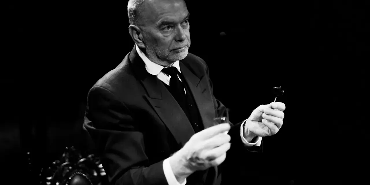 Piotr Wyszomirski w eleganckim garniturze, trzymający w dłoniach kieliszek i coś na patyczku.