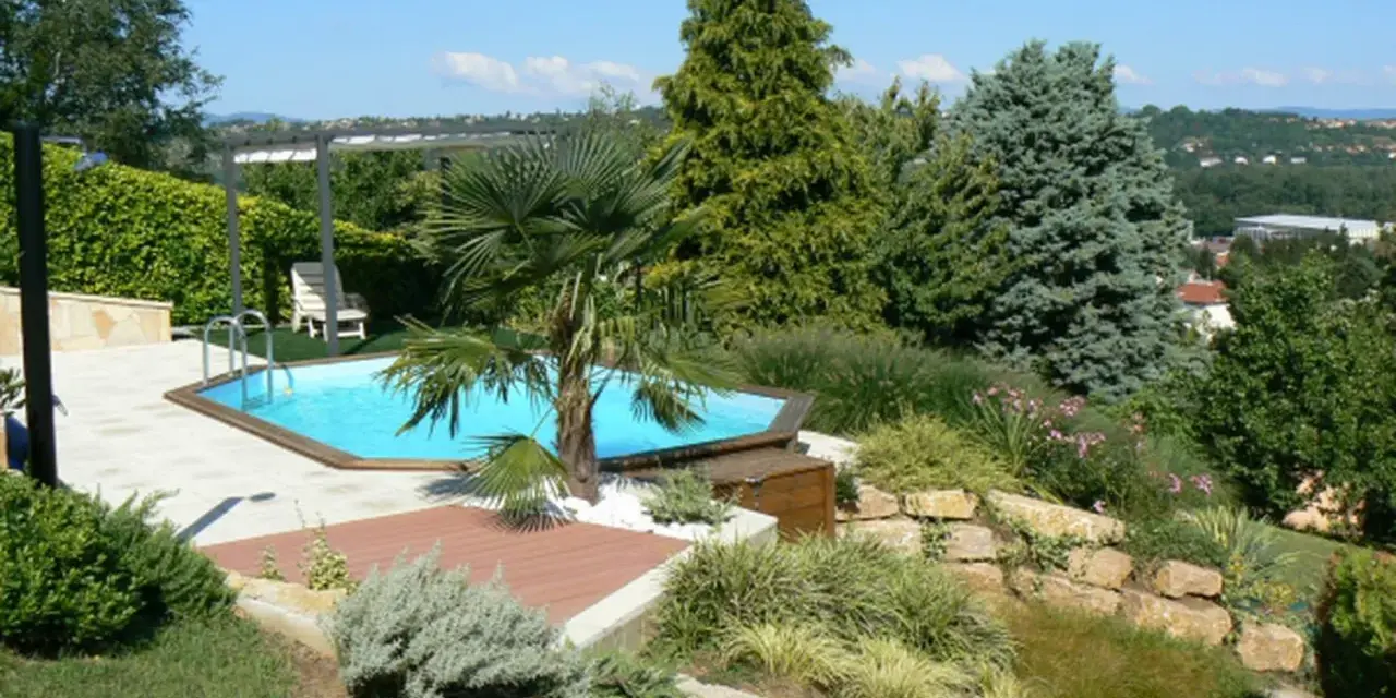 Piscine aux formes douces sur un terrain en pente, entourée de végétation luxuriante et d'une terrasse en bois.