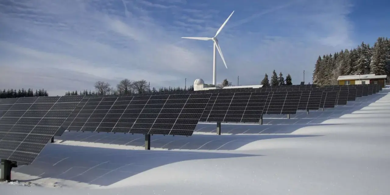 Rzędy paneli bifacjalnych w śniegu, z wiatrakiem i budynkiem w tle.