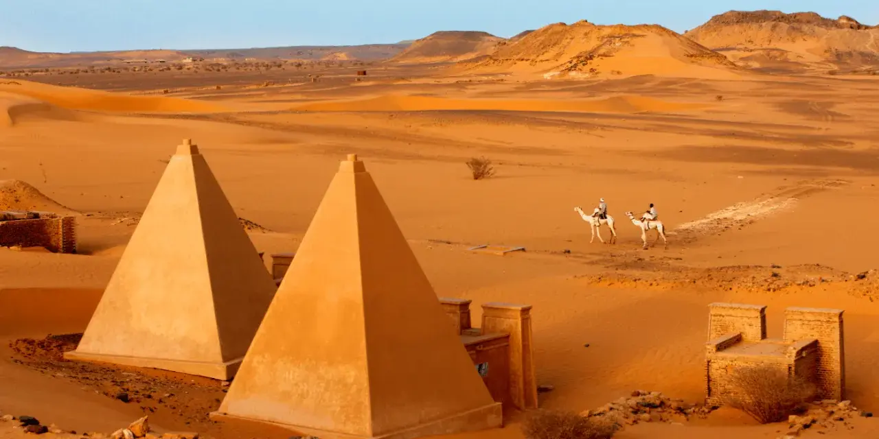 Pustynny krajobraz Sudanu z piramidami i dwoma podróżnikami na wielbłądach.