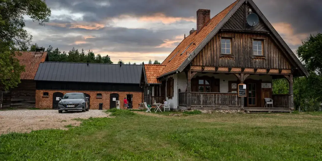 Szelągówka camping: drewniane domki i budynki gospodarcze, samochód na żwirowym podjeździe, zachmurzone niebo.