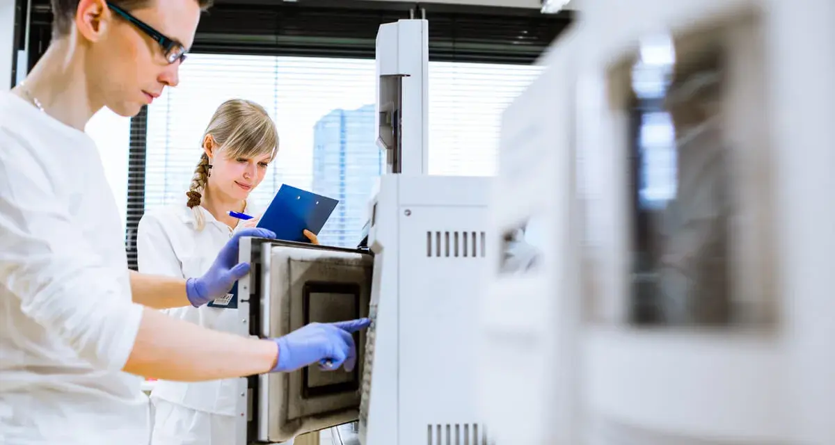 W laboratorium młoda para naukowców pracuje przy aparaturze. On w okularach, ona z notatkami.