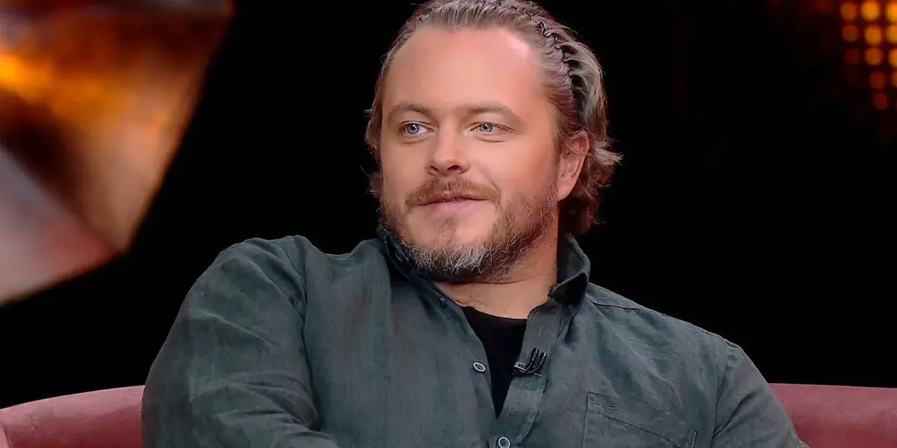 Tomasz Schuchardt, znany z wielu filmów, seriali i programów, z charakterystyczną fryzurą i niebieskimi oczami, siedzi na kanapie.