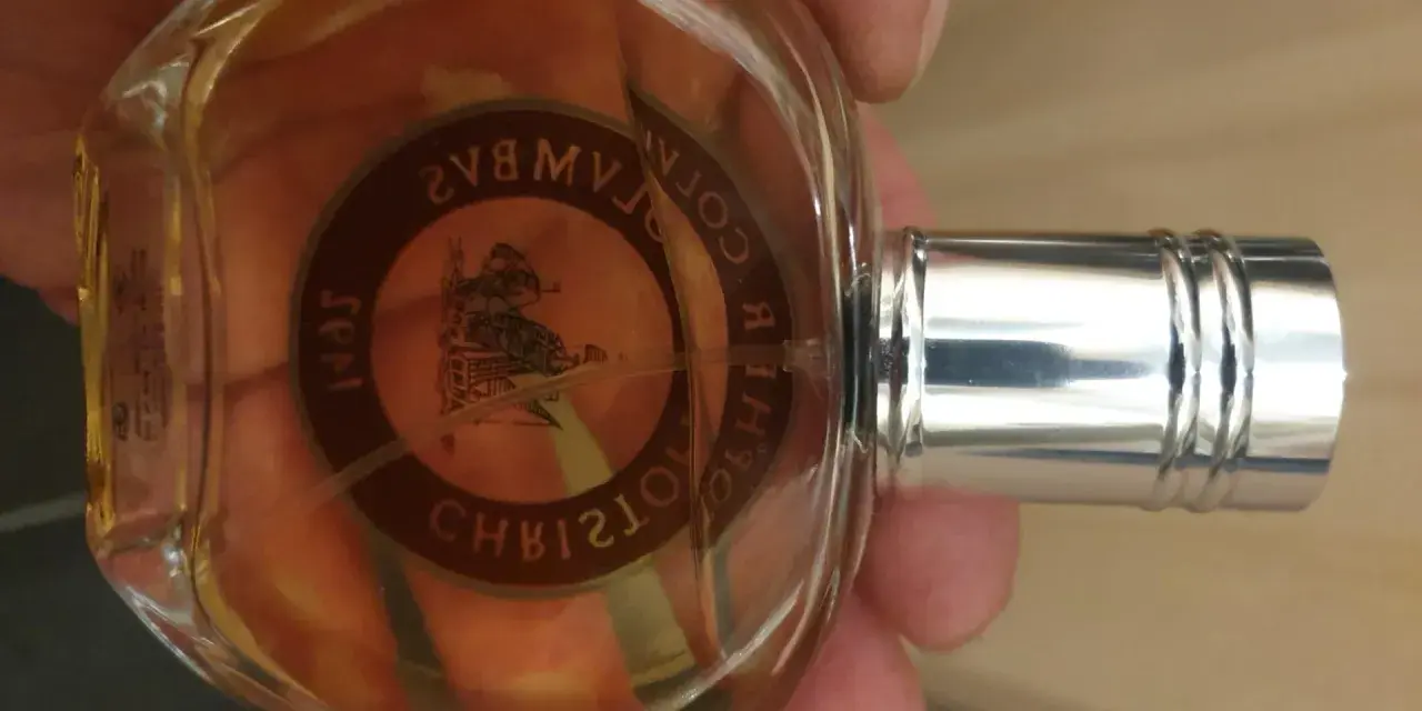 Szklana butelka z bursztynowym płynem, ozdobiona okrągłą etykietą z wizerunkiem statku i napisem "Christopher Columbus perfumy".
