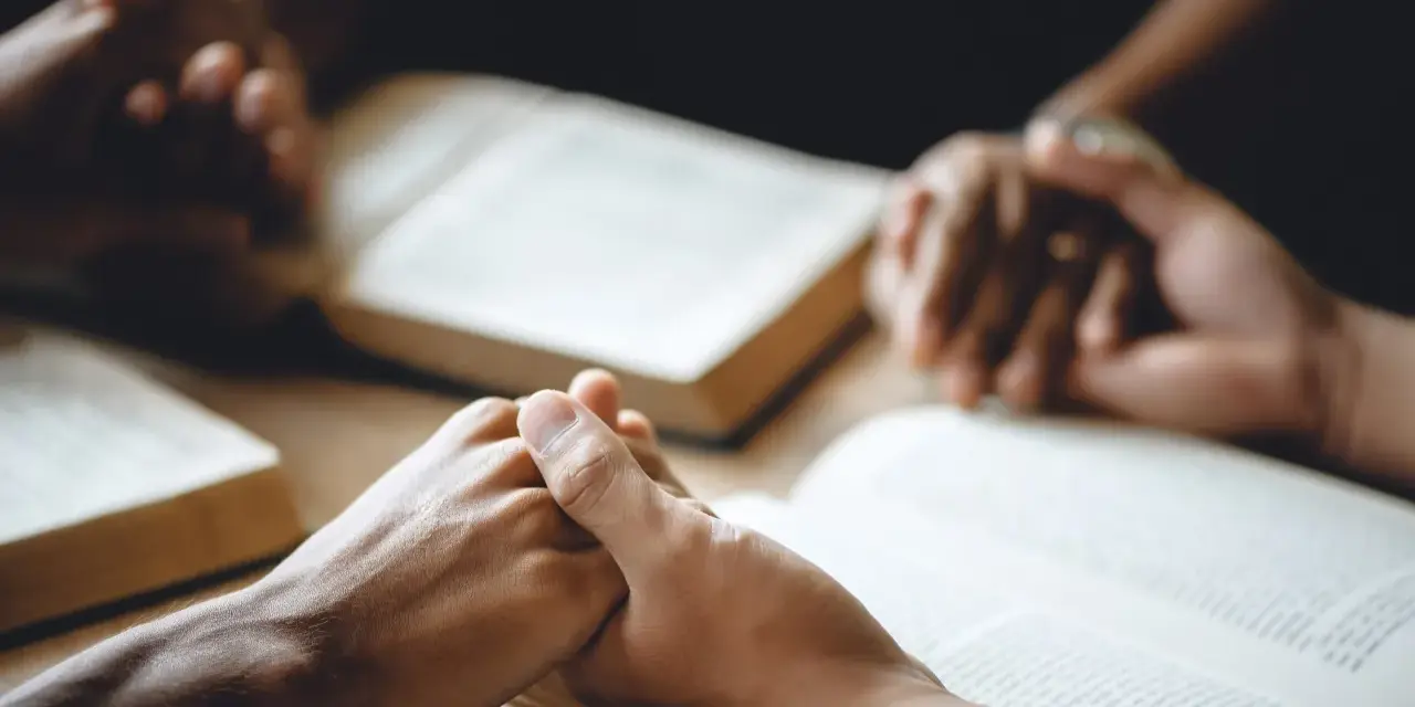 Hände halten sich über aufgeschlagenen Bibeln, ein Zeichen dafür, wie beten Christen: in Gemeinschaft, mit Glauben und Hoffnung.