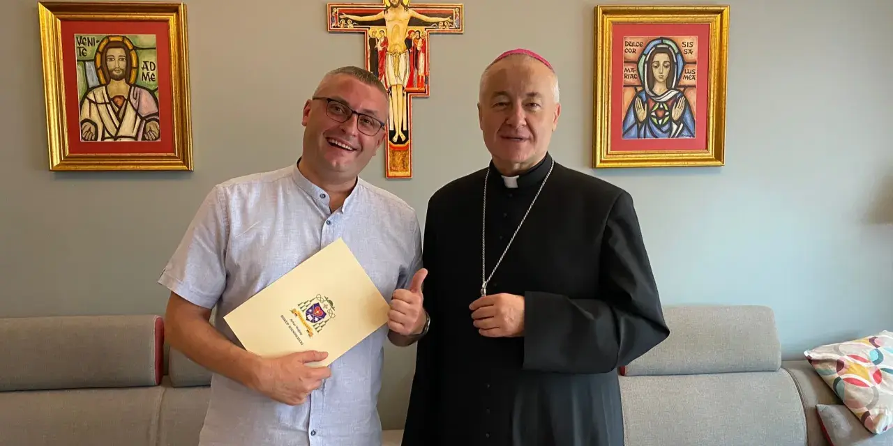 Uśmiechnięty mężczyzna z dokumentem i biskup pozują na tle krzyża i obrazów. Mężczyzna trzyma dokument z herbem, a biskup ma na sobie sutannę.