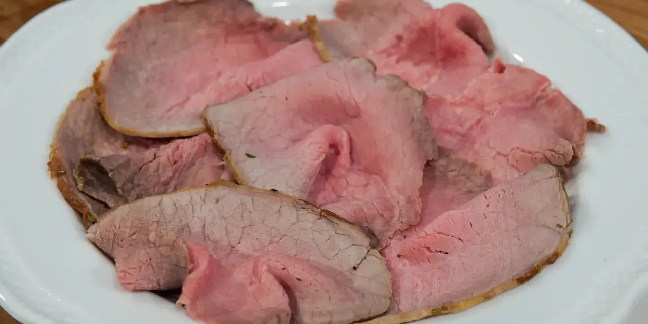 Fette sottili di arrosto di manzo rosa e succulento, servite su un piatto bianco.