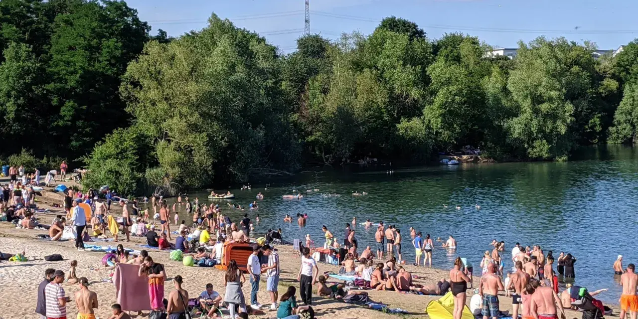 Ein sonniger Tag am **Badesee Bonn**. Viele Menschen genießen das Wasser und den Strand, umgeben von grünen Bäumen.