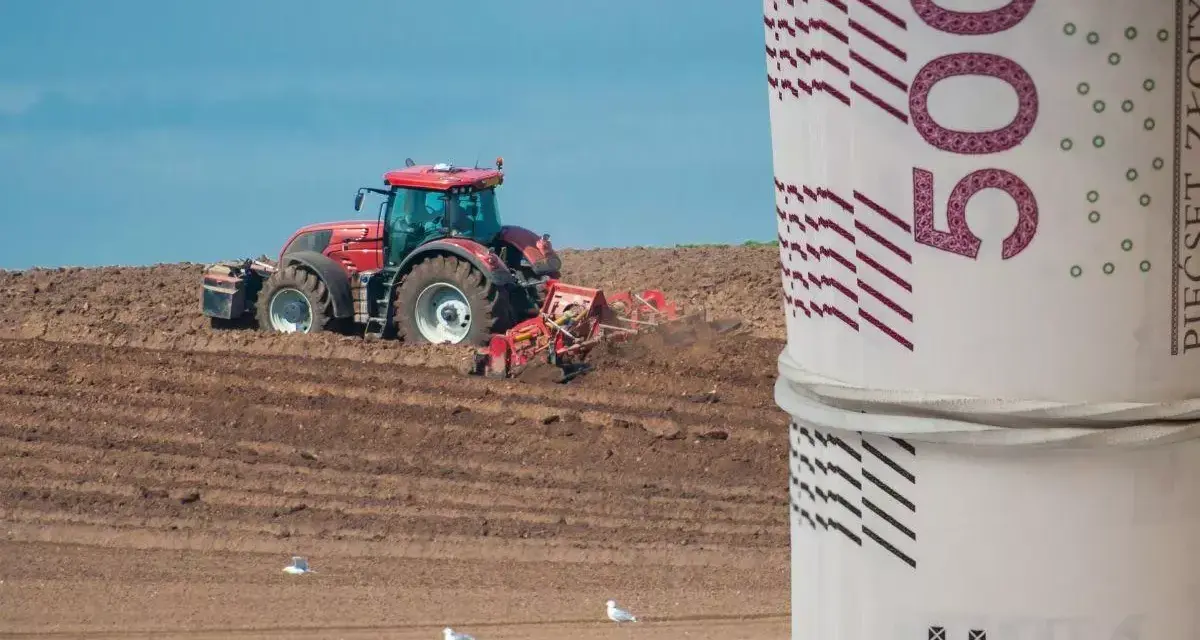 Czerwony traktor orze pole, a na pierwszym planie zwinięte banknoty 500 zł. Zastanawiasz się, ile kosztuje hektar ziemi budowlanej?