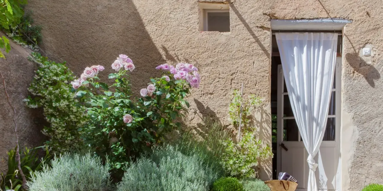 Lavendel beet ideen: Ein mediterraner Garten mit Rosen, Lavendel und einer Tür mit weißem Vorhang.