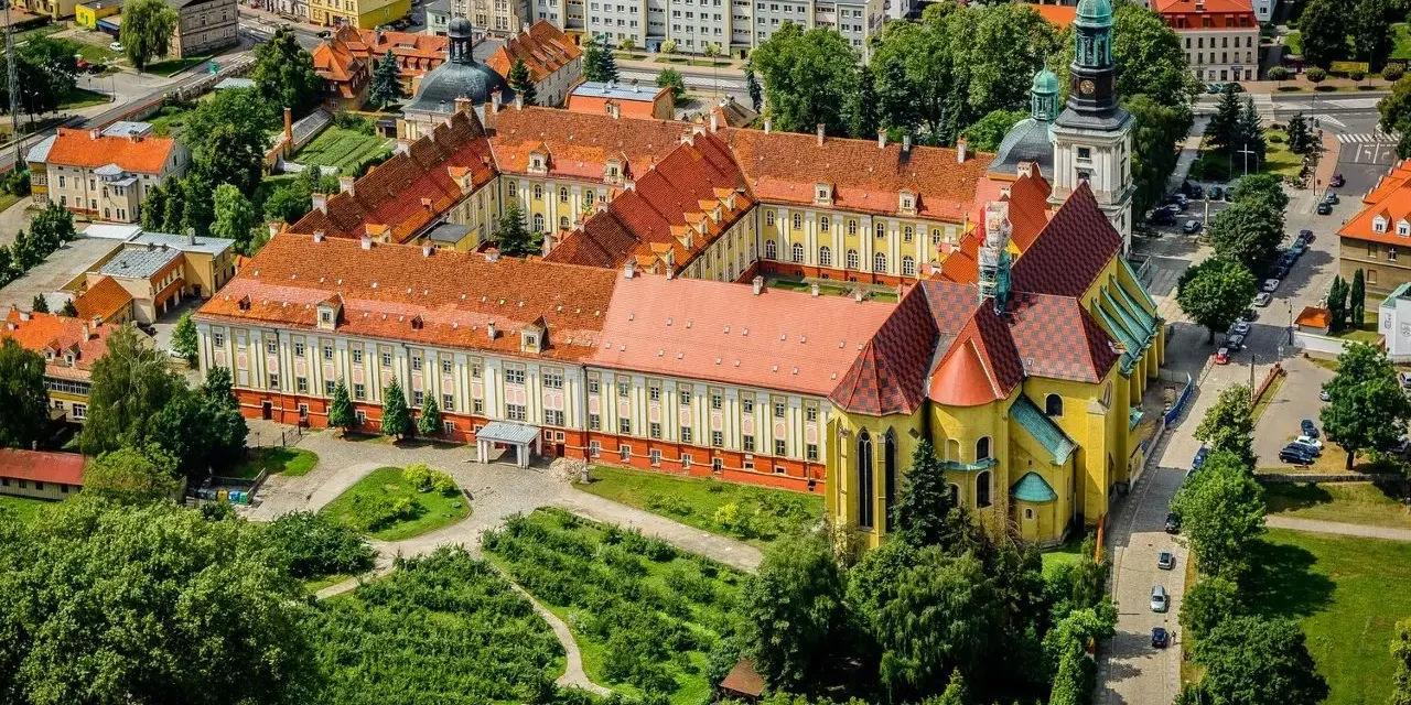 Widok z lotu ptaka na klasztor w Trzebnicy z charakterystycznymi żółtymi murami i czerwonymi dachami. Otoczony zielenią i budynkami miejskimi.