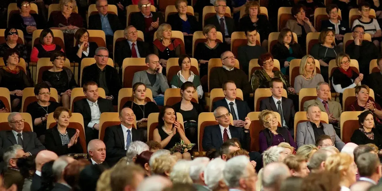 Elegancko ubrani ludzie siedzą w filharmonii. Panie w sukienkach, panowie w garniturach. To inspiracja, jak się ubrać do filharmonii.