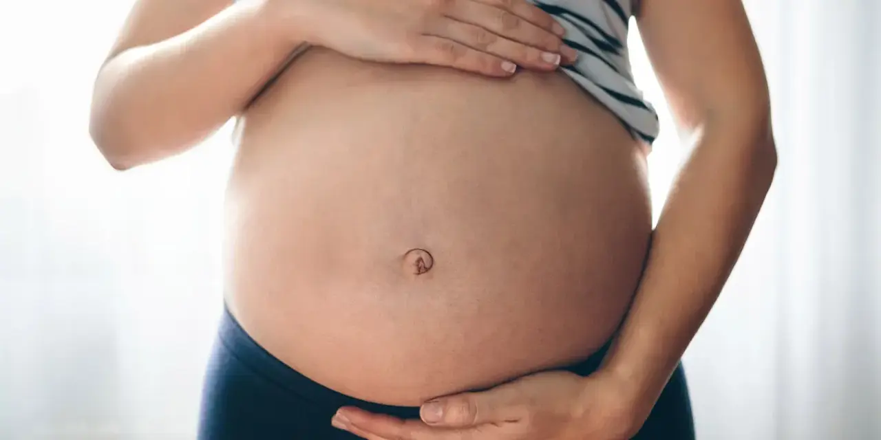 Brzuszek w ciąży z widocznym pępkiem, otoczony dłońmi przyszłej mamy.