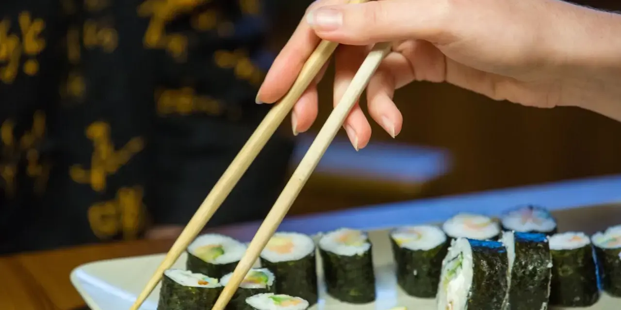 Dłoń z pałeczkami sięga po rolkę sushi. Na talerzu ułożone są kawałki sushi, każdy z nich to mała porcja smaku i kalorii.