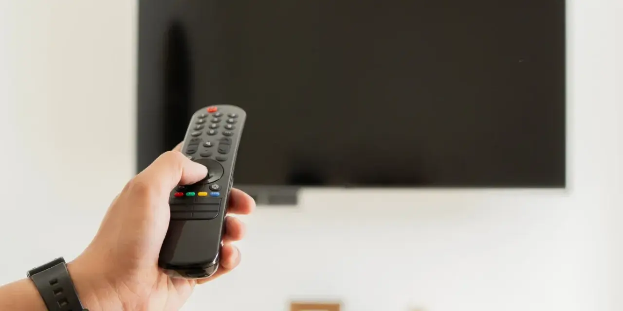Dłoń trzymająca pilot do telewizora na tle wyłączonego ekranu.