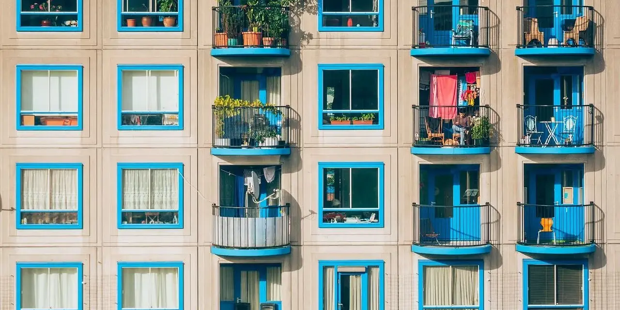Fachada de edificio con ventanas y balcones de marcos azules. Cada balcón muestra detalles de la vida cotidiana, como plantas y ropa tendida, reflejando los gastos de comunidad.