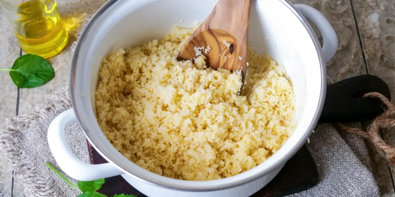 Ein Holzlöffel lockert den frisch zubereiteten Couscous in einem Topf auf. Daneben stehen Öl und Minze.