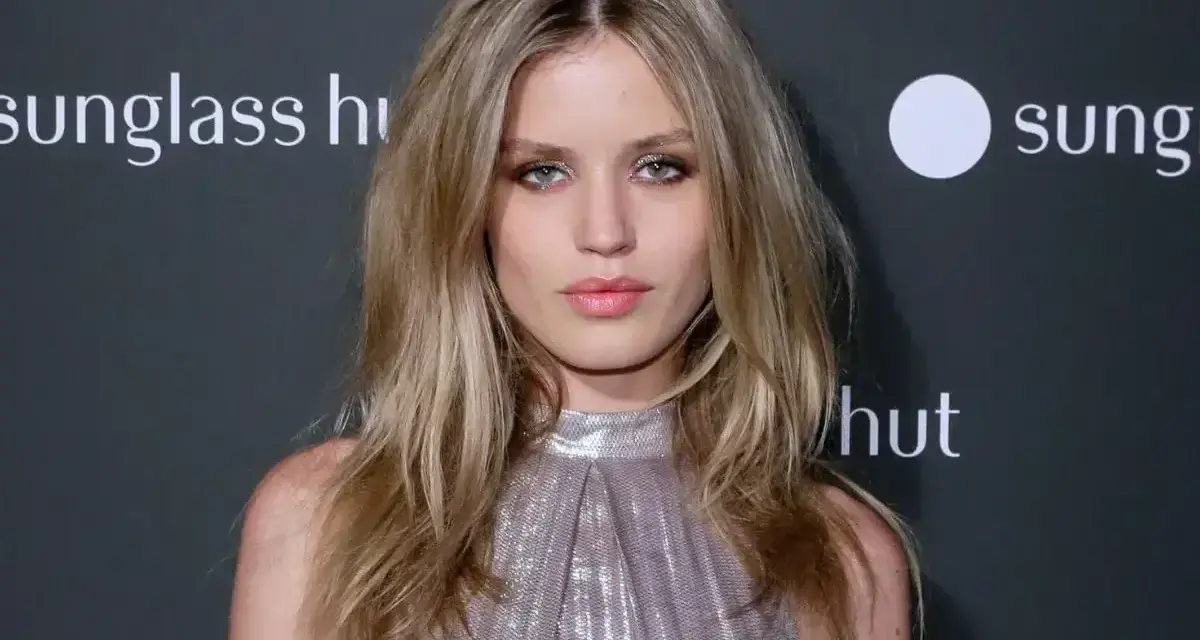 Modelka z chłodnymi odcieniami blondu, w błyszczącej sukience, pozuje na tle logo Sunglass Hut.