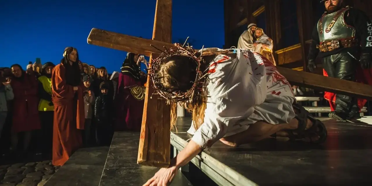 Scena drogi krzyżowej: Jezus w cierniowej koronie, z krwią na szacie, upada pod ciężarem krzyża. Tłum widzów obserwuje to poruszające przedstawienie.