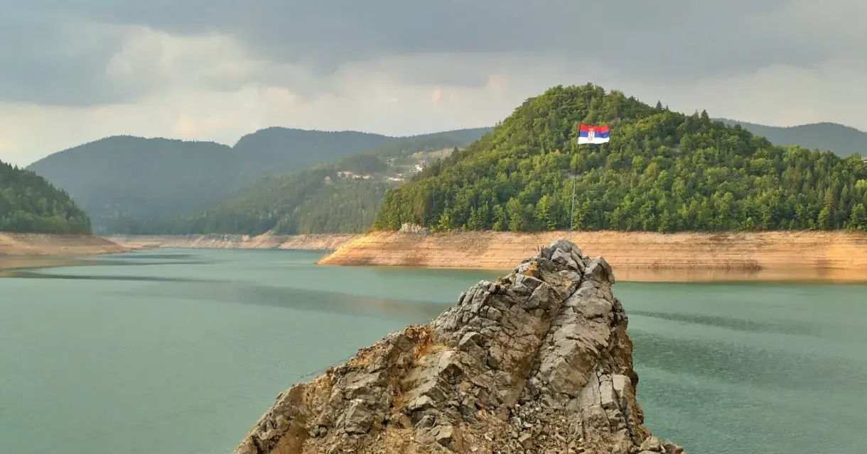Serbska flaga na skale nad jeziorem. Krajobraz zachęca do podróży, a pytanie "czy Serbia jest bezpieczna dla turystów" wydaje się nie mieć znaczenia w obliczu tego spokoju.