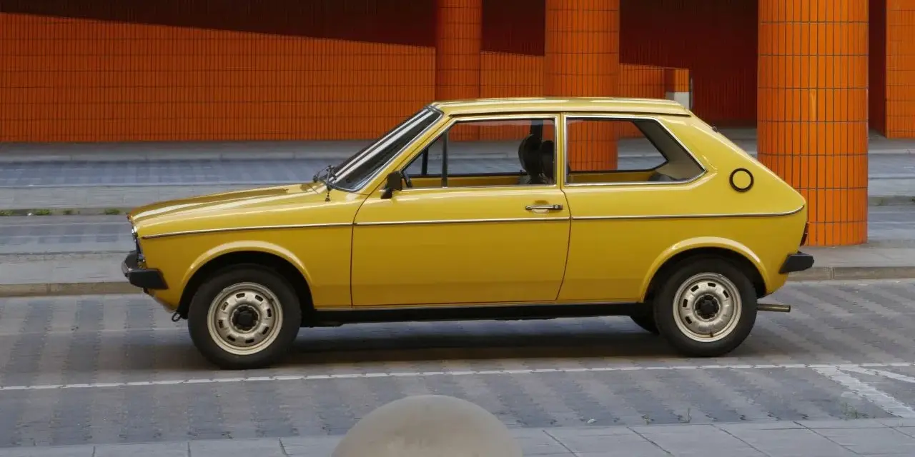 Żółty Audi 50, klasyk z lat 70., parkuje na tle pomarańczowej, kafelkowej architektury.