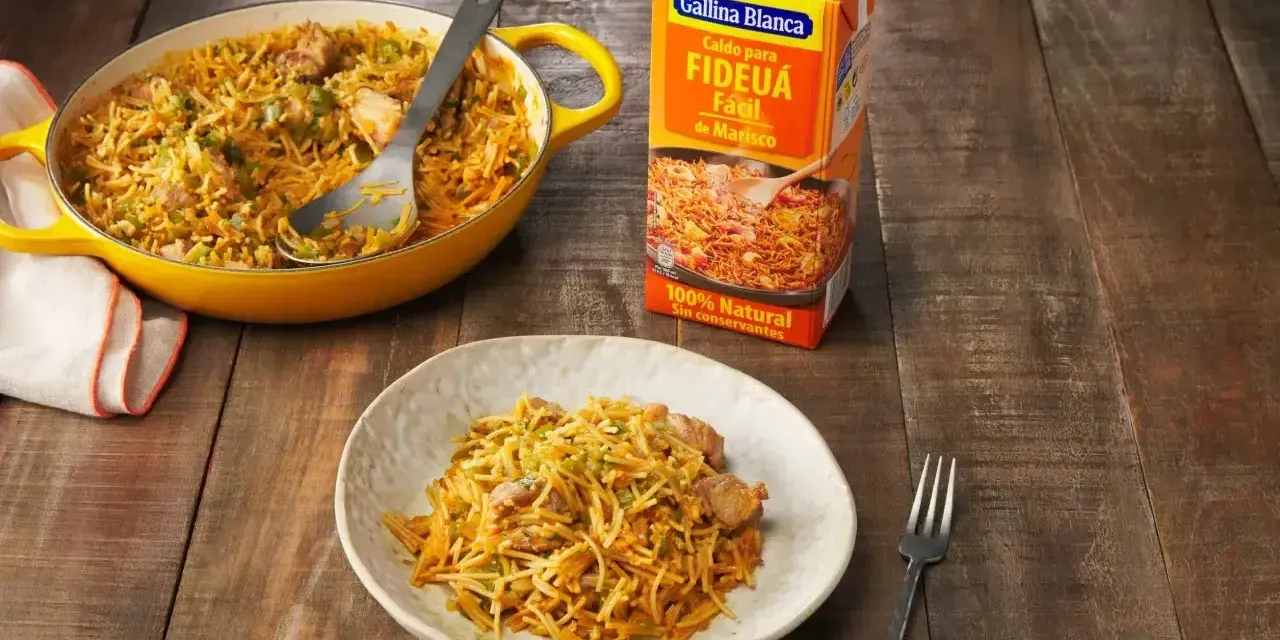 Un plato de fideos a la cazuela servido en una cazuela amarilla y en un plato blanco, junto a un cartón de caldo para fideuá.