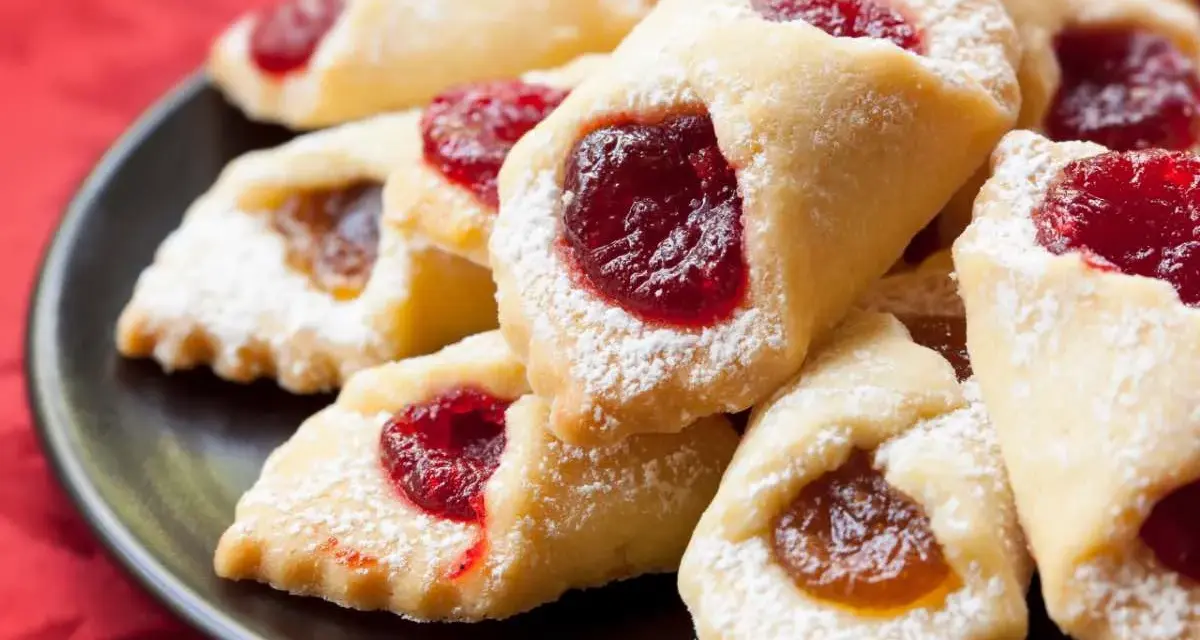 Ciastka kruche con ripieno di marmellata, spolverati di zucchero a velo, su un piatto scuro.