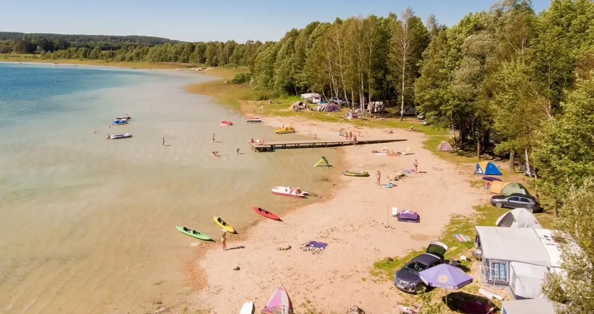Letnie popołudnie nad jeziorem Świętajno. Ludzie kąpią się, pływają kajakami i odpoczywają na plaży wśród namiotów i kamperów.