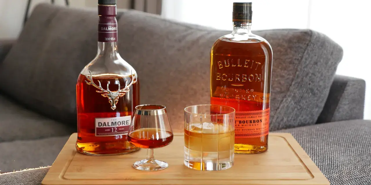 Zwei Flaschen edler Tropfen: Dalmore Scotch und Bulleit Bourbon. Ein Glas Scotch und ein Glas Bourbon mit Eis stehen bereit.