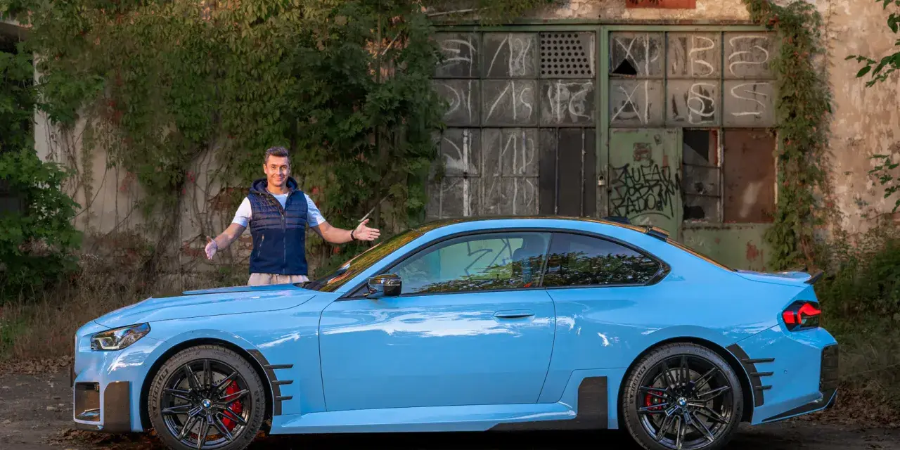 Mężczyzna prezentuje błękitne BMW M2 G87 przed zniszczonym budynkiem.