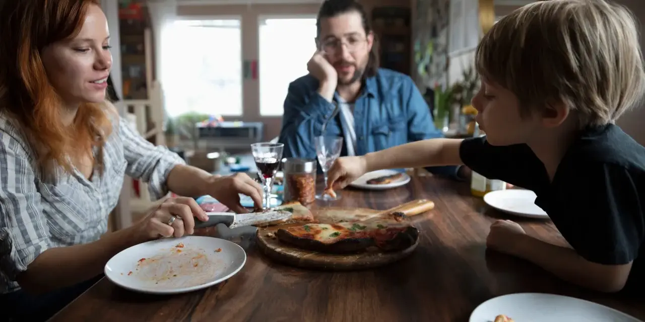 Rodzina przy stole, pizza, w tle mężczyzna zamyślony. Czasem nawet przy wspólnym posiłku pojawiają się kłótnie o dzieci z poprzedniego związku.