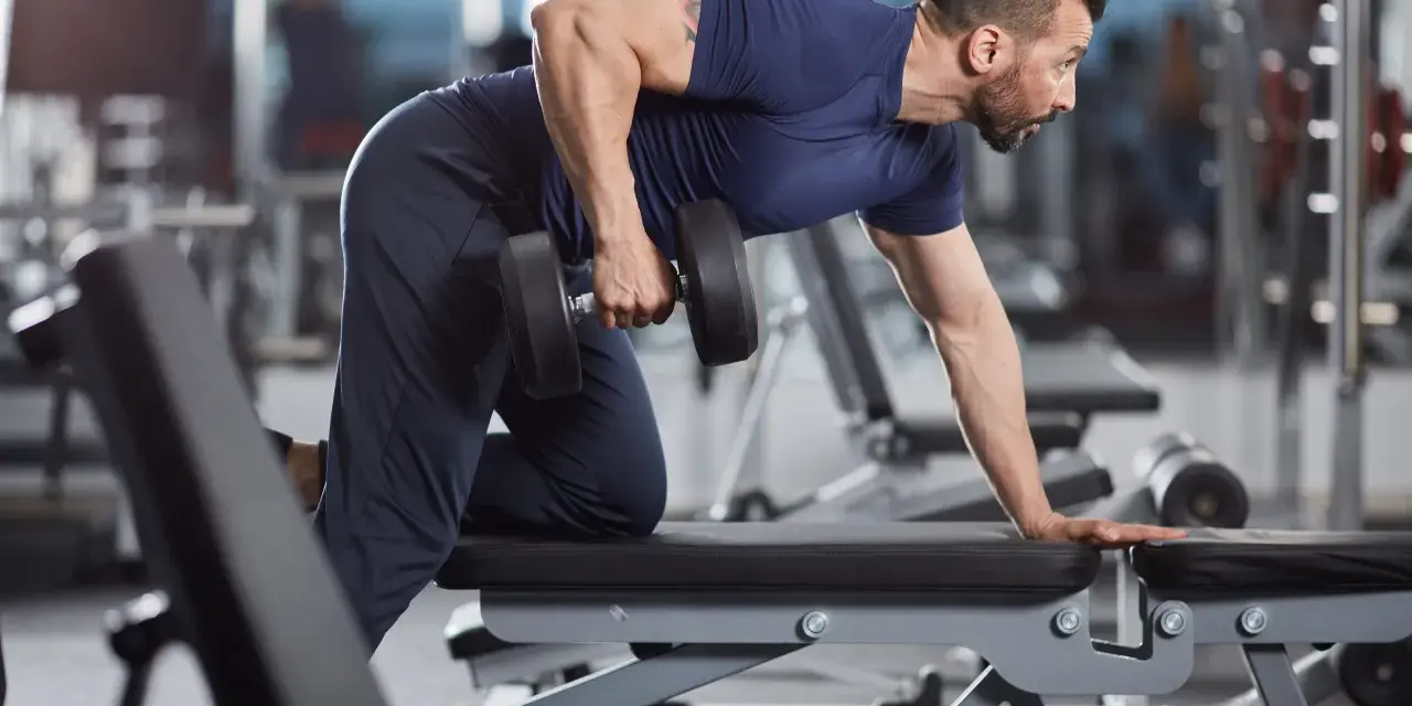 Man utför enarms hantelrodd på ett gym. Han stöttar sig mot en bänk med ena handen och drar en hantel mot bröstet med den andra.