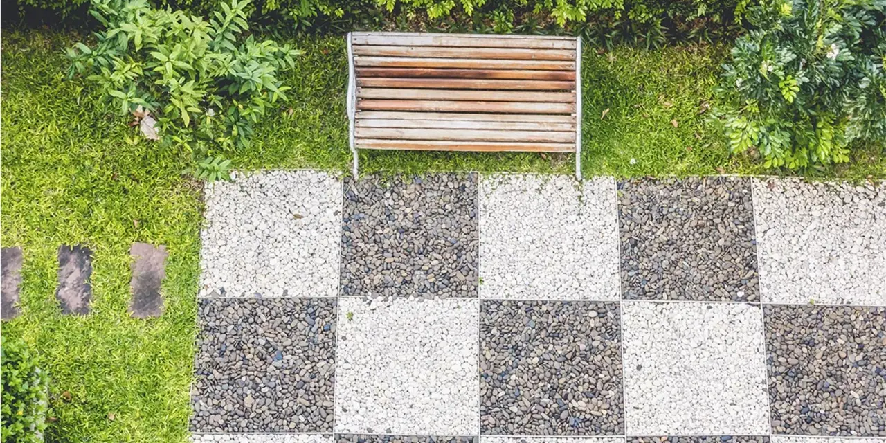 Ideas de suelo para terraza: un banco de madera sobre césped y un patrón de tablero de ajedrez de guijarros blancos y grises.