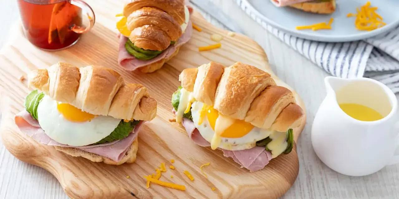 Trois croissants au jambon, œuf au plat, avocat et fromage râpé, servis sur une planche en bois avec une tasse de thé.