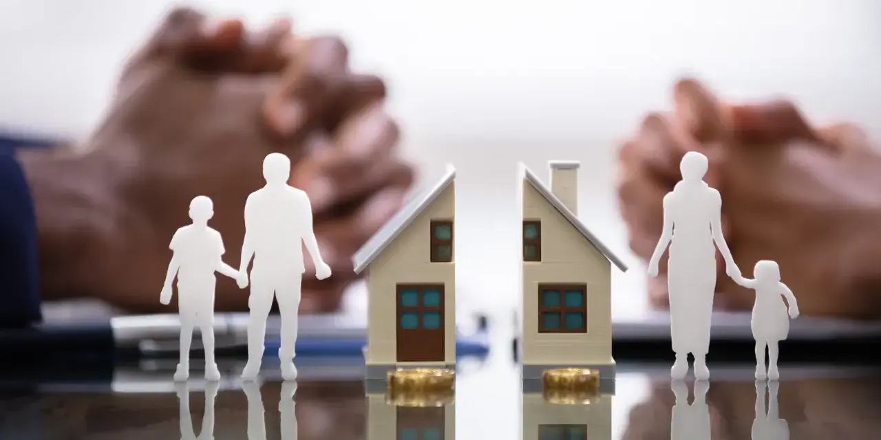 Figurines de famille séparées par une maison coupée en deux, symbolisant une convention d'indivision divorce. Mains floues en arrière-plan.