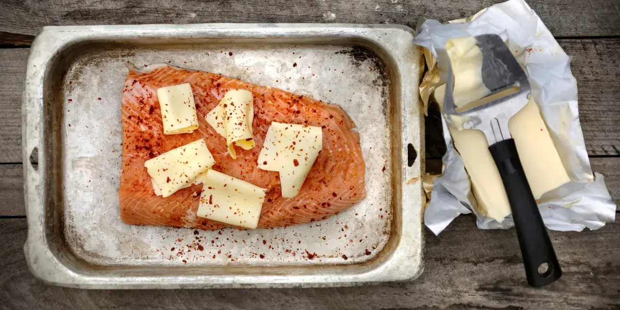 Lax i ugn, kryddad med chili och toppad med ostskivor, redo att tillagas. Bredvid ligger smör och en osthyvel.